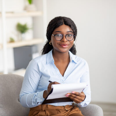 Friendly black female psychologist writing in clipboard, having session with client, sitting on couch at modern office. Happy psychotherapist taking notes during consultation in mental health clinic